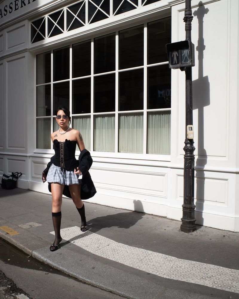 Une invitée en bustier noir et jupe courte marche sur le trottoir devant une façade blanche, à Paris